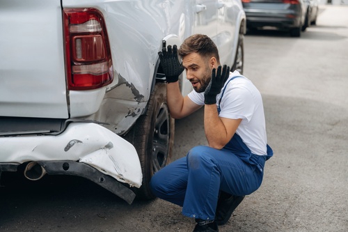mechanic in overalls crouching beside vehicle with rear bumper collision damage