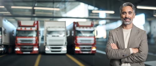 confident businessman standing arms crossed before fleet of commercial semi-trucks